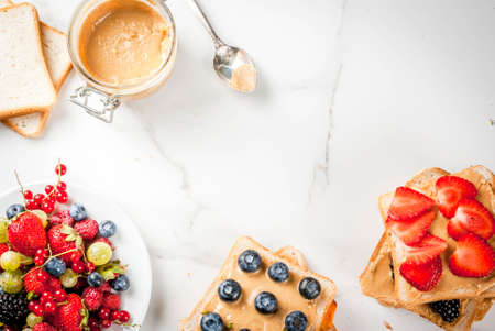 Traditional American and European summer breakfast: sandwiches of toast with peanut butter, berry, fruit apple, peach, blueberry, blueberry, strawberry, banana. White marble table. copy space top viewの写真素材