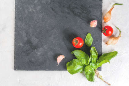 Food background. Ingredients, greens and spices for cooking lunch, lunch. Fresh basil leaves, tomatoes, garlic, onions, salt, pepper. On a white stone table, a slate board. Top view copy spaceの写真素材
