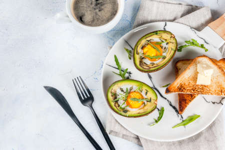 Healthy vegan breakfast. Diet. Baked avocado with egg and fresh salad from arugula, toast and butter. On a white marble plate, a light concrete table. A cup of coffee. Copy space top viewの写真素材