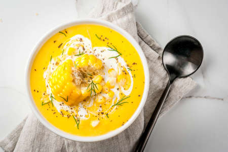 Vegan cuisine. Traditional autumn corn soup. Served with corn cobs, greens, cream. On a white marble table. Copy space top viewの写真素材