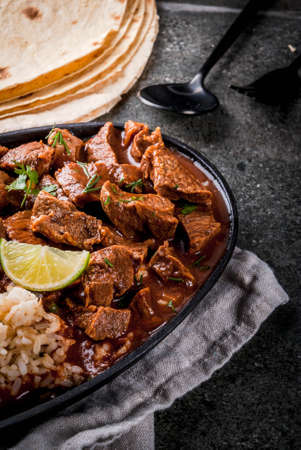 Mexican and American traditional food. Stew beef with tomatoes, spices, pepper - Chile Colorado. With boiled rice. lime, tortillas.  Copy space. On black stone tableの写真素材