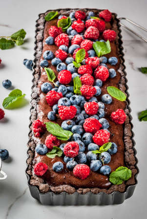 Summer homemade baked pastry. Chocolate cake tart with chocolate cream, fresh raw berries blueberry raspberry, decorated with mint leaves, powdered sugar. On white marble table, copy spaceの写真素材