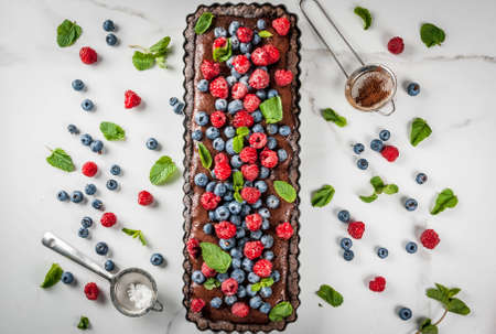 Summer homemade baked pastry. Chocolate cake tart with chocolate cream, fresh raw berries blueberry raspberry, decorated with mint leaves, powdered sugar. On white marble table, copy space top viewの写真素材
