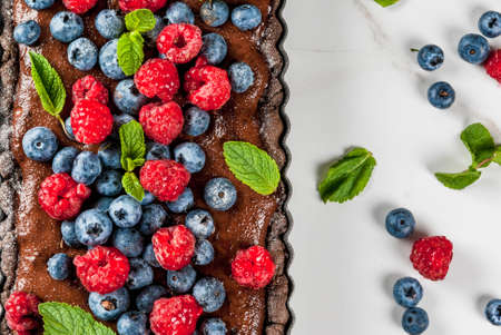 Summer homemade baked pastry. Chocolate cake tart with chocolate cream, fresh raw berries blueberry raspberry, with mint leaves, powdered sugar. White marble table, close top view copy spaceの写真素材