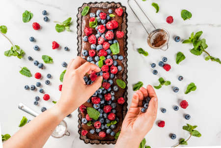 Summer homemade baked pastry. Chocolate cake tart with chocolate cream, woman decorate it with blueberry raspberry mint leaves, powdered sugar. White marble table, copy space top view hands in pictureの写真素材