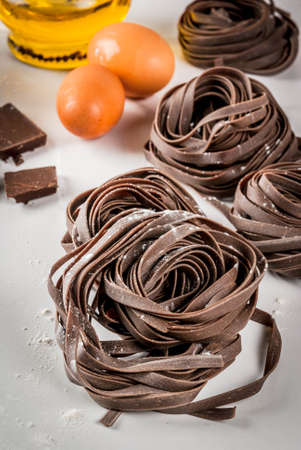 Raw unprepared chocolate pasta noodles, with ingredients for cooking - chocolate, cocoa, flour, eggs,  oil. On a white kitchen marble table. Copy spaceの写真素材