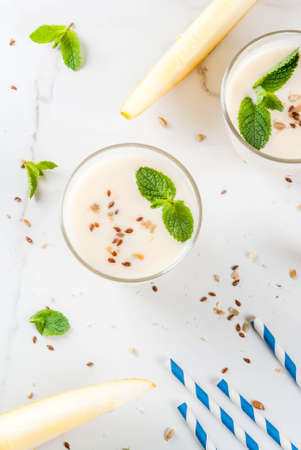 Summer fruits, refreshing drinks. Vegan food. Breakfast. Smoothie of yogurt, raw organic yellow melon, flax seeds and mint. With striped straws, in glasses, white marble table, copy space  top viewの写真素材