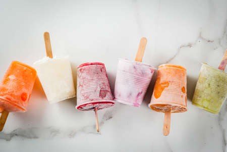 Healthy summer desserts. Ice cream popsicles. Frozen tropical juices, smoothies blueberries. currants, orange, mango, kiwi, banana, coconut, raspberry. On white marble table copy space top viewの写真素材