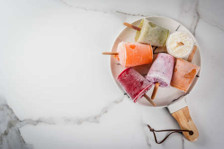 Healthy summer desserts. Ice cream popsicles. Frozen tropical juices, smoothies blueberries. currants, orange, mango, kiwi, banana, coconut, raspberry. On white marble table, plate copy space top viewの写真素材