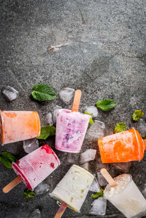 Healthy summer desserts. Ice cream popsicles. Frozen tropical juices, smoothies blueberries. currants, orange, mango, kiwi, banana, coconut, raspberry. On black stone table, plate copy space top viewの写真素材