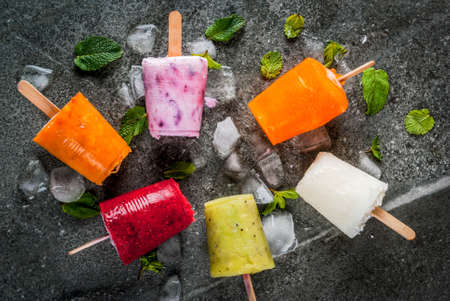Healthy summer desserts. Ice cream popsicles. Frozen tropical juices, smoothies blueberries. currants, orange, mango, kiwi, banana, coconut, raspberry. On black stone table copy space top viewの写真素材