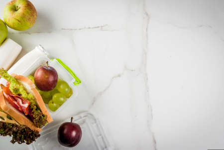 Back to school. A healthy lunch in a box is fresh fruit (apples, plums, grapes), a bottle of yogurt and a sandwich with lettuce, tomatoes, cheese, meat. White marble table. Copy space top viewの写真素材