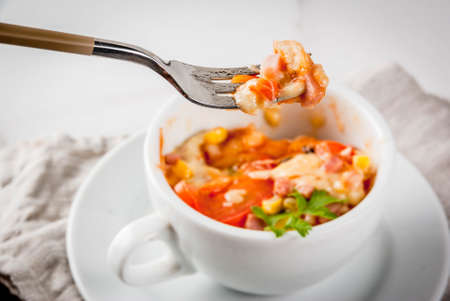 Trendy modern food, fast food, lunch in the microwave. Pizza in a mug, with ham, tomatoes, mushrooms, cheese. White cup, with a fork, on a white marble table. Copy spaceの写真素材