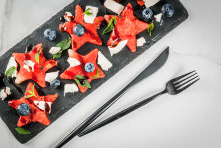 Summer fruit berry salad: watermelon sliced in the form of stars, blueberries, feta cheese and balsamic sauce. On a black slate, on a white marble table. With a fork and knife. Top view copy spaceの写真素材