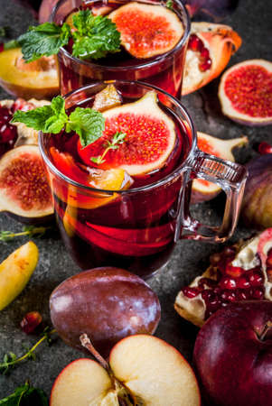 Warming autumn, winter cocktail drinks recipes. Hot red fruit sangria with apples, plums, figs, pomegranate, mint, cinnamon, thyme, lemon. On dark stone table, copy spaceの写真素材