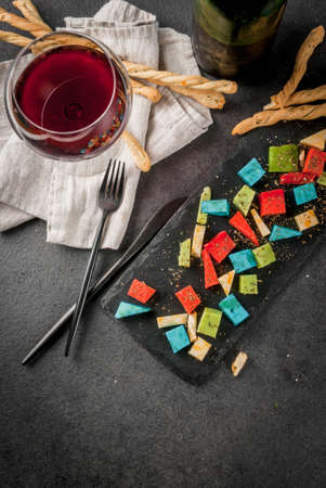 Trendy modern food. Multicolored organic holland cheese - blue (lavender), red (hot pepper), green (basil), classic yellow cheese. With bread sticks, wine glass, black table. Copy space top viewの写真素材