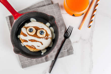 Funny food for Halloween. Kids breakfast pancake decorated like mummy with white chocolate sauce, banana, berries, with pumpkin smoothie juice, white table copy space top viewの写真素材