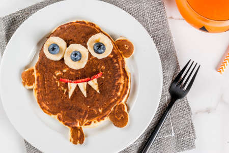 Funny food for Halloween. Kids breakfast pancake decorated like creepy monster, with banana, berries, with pumpkin smoothie juice, white table copy space top viewの写真素材