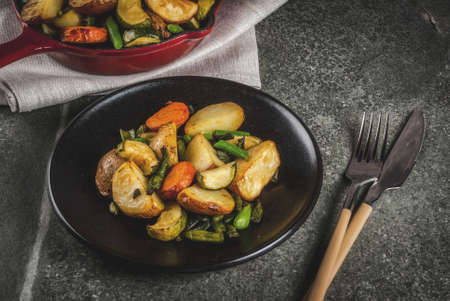 Plate and Skillet with  fried seasonal autumn vegetables (zucchini, potatoes, carrots, beans), on black stone table copy spaceの写真素材