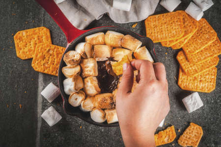 Indoor sâmores, baked sâmores dip in a cast iron skillet pan with graham crackers. Girls hand in picture, tasting smores, dark grey table, top view copy spaceの写真素材