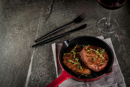 Lunch for one person. Homemade grilled beef steaks with thyme in a portioned frying pan, with a fork, knife and a glass of wine.  On black stone table, copy space top viewの写真素材