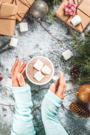 Christmas concept, table background with snow, christmas tree branches, girl holds hot chocolate mug wit marshmallow, gifts or presents, pine cones and decoration, copy space top viewの写真素材