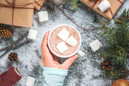 Christmas concept, table background with snow, christmas tree branches, girl holds hot chocolate mug wit marshmallow, gifts or presents, pine cones and decoration, copy space top viewの写真素材