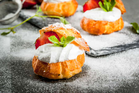 Summer desserts. Homemade baking. Cake profiteroles with whipped cream, fresh strawberries, mint and sprinkling of powdered sugar. On a black stone table. Copy spaceの写真素材