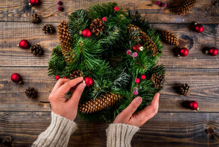 Preparation for xmas holidays. Woman decorating christmas green wreath with pine cones and red winter berries and christmas tree balls, wooden background, top view copy space, female hands in pictureの写真素材
