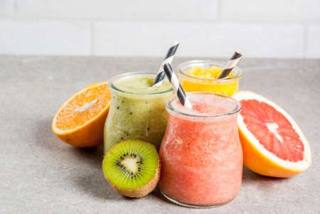 Detox organic diet drinks, homemade tropical smoothies - kiwi, orange, grapefruit, in portioned jars, with ingredients, on a gray stone table. Copy spaceの写真素材