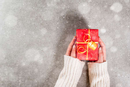 Preparing for the holidays, Christmas background. Female hands in the picture in a warm sweater hold a gift in a red wrapper with a golden ribbon. Gray background, snow effect, Top view copy spaceの写真素材