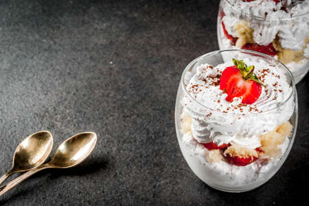 Layered dessert parfait in a glass with strawberries, sponge cake and whipped cream. On a black stone table. Copy spaceの写真素材