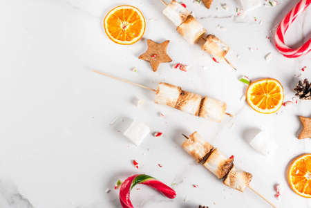 Traditional christmas sweets - candy cane, marshmallow, dried orange, gingerbread stars, baked on fire marshmallow skewers on white marble background, copy space top viewの写真素材