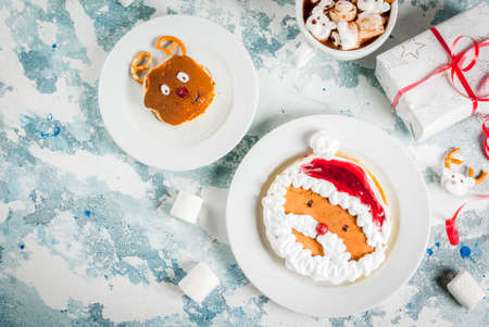 Idea for a children's Christmas breakfast: pancakes decorated like Santa Claus and deer, cocoa with teddy bears and deer marshmallow. On a light blue background, with xmas gift top view copy spaceの写真素材