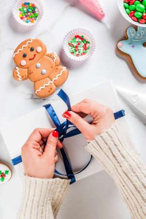Preparing for Christmas, decorating traditional gingerbread with multicolored sugar glaze, the girl folds the cookies in a white gift box, with ribbon bow, on a white marble table copy space top viewの写真素材