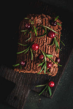 Traditional Christmas dessert, Christmas yule log cake with chocolate cream, cranberry and rosemary twigs. On a dark background with Christmas tree branches, gifts and decorations, copy space top viewの写真素材