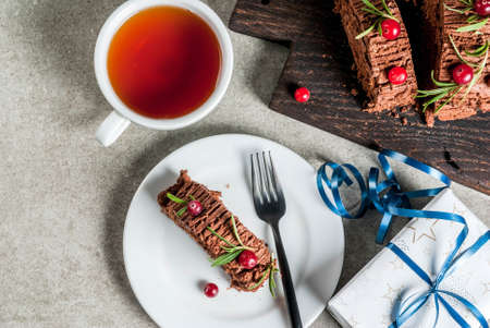 Traditional Christmas dessert, Christmas yule log cake with chocolate cream, cranberry and rosemary twigs. On stone gray background with Christmas gifts and tea, copy space top viewの写真素材