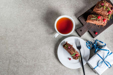 Traditional Christmas dessert, Christmas yule log cake with chocolate cream, cranberry and rosemary twigs. On stone gray background with Christmas gifts and tea, copy space top viewの写真素材