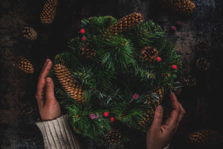Preparation for xmas holidays. Woman decorating christmas green wreath with pine cones and red winter berries, on dark rusty  background, top view copy space, female hands in pictureの写真素材