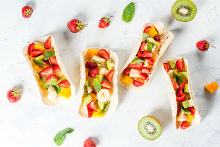 Summer snacks. Food for a party. Fruit tacos with strawberries, mangoes, bananas, chocolate, mint. On a light blue concrete table. Copy space top viewの写真素材