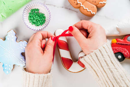 Preparing for Christmas, decorating traditional gingerbread with multicolored sugar icing, white marble table. Girl (hands in picture) knot ribbon bow on candy cane gingerbread, top view copy spaceの写真素材