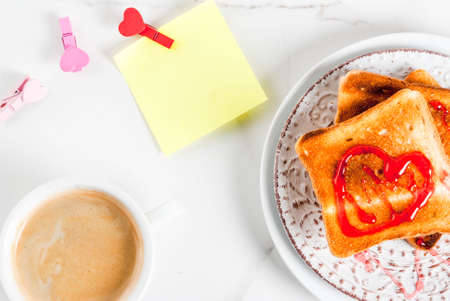 Valentine's day breakfast idea with coffee mug, toasted bread with red strawberry jam, blank paper note for congratulations with heart shaped pins, white marble background, copy space top viewの写真素材