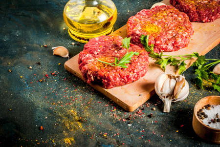 Raw burger cutlets with salt, pepper, oil, herbs and spice, on dark table, copy spaceの写真素材