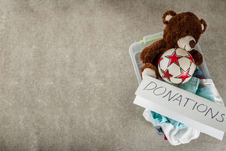 Plastic donation box with toys, clothes and food, white grey background copy space top view