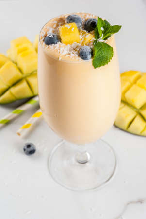 Dietary drink, breakfast. Tropical mango smoothie with fresh pieces of mango, blueberries, coconut and mint leaves. In a glass, on a white marble table. Copy spaceの写真素材