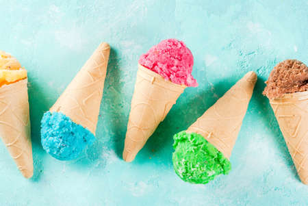 Selection of various bright multicolored ice-cream in ice cream cones - chocolate vanilla blueberry strawberry pistachio orange, on light blue sunny background, copy space top viewの写真素材