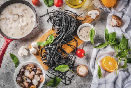 Raw uncooked black cuttlefish ink spaghetti pasta with ingredients for cooking dinner - olive oil, basil, tomatoes, champignons, lemon, seafoods,, on grey stone table top viewの写真素材