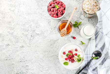 Raw ingredients for summer healthy breakfast, cereals (oats), fresh Raspberry, mint leaves, yoghurt, honey, on light concrete background, copy space top viewの写真素材