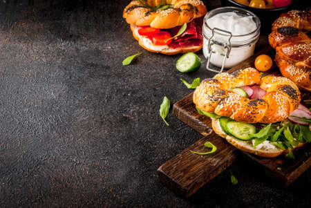 Variety of homemade bagels sandwiches with sesame and poppy seeds, cream cheese,  ham, radish, arugula, cherry tomatoes, cucumbers, with ingredients on dark concrete background copy spaceの写真素材