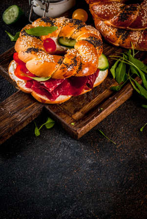 Variety of homemade bagels sandwiches with sesame and poppy seeds, cream cheese,  ham, radish, arugula, cherry tomatoes, cucumbers, with ingredients on dark concrete background copy spaceの写真素材